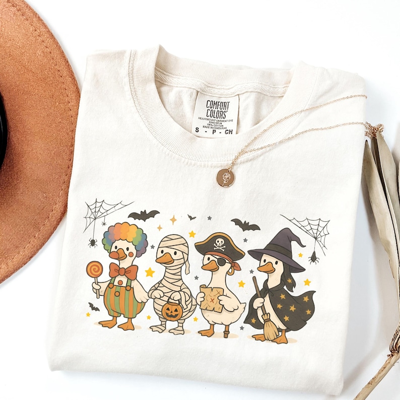 May include: Cream-colored t-shirt featuring a Halloween-themed graphic of four ducks dressed in costumes: a clown, a mummy, a pirate, and a witch. The shirt includes a Comfort Colors tag and a gold necklace. The design includes bats, spiders, and stars.