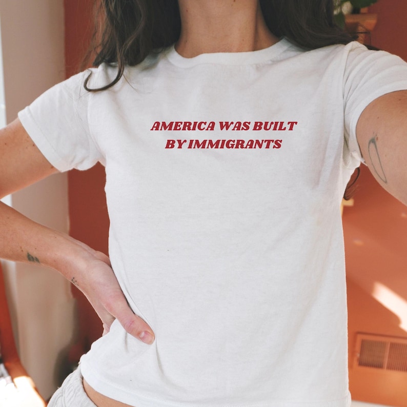 White baby tee with y2k style red text reading "America Was Built By Immigrants." A pro-immigration shirt and political activist t-shirt for equal rights supporters. Perfect progressive gift for her.