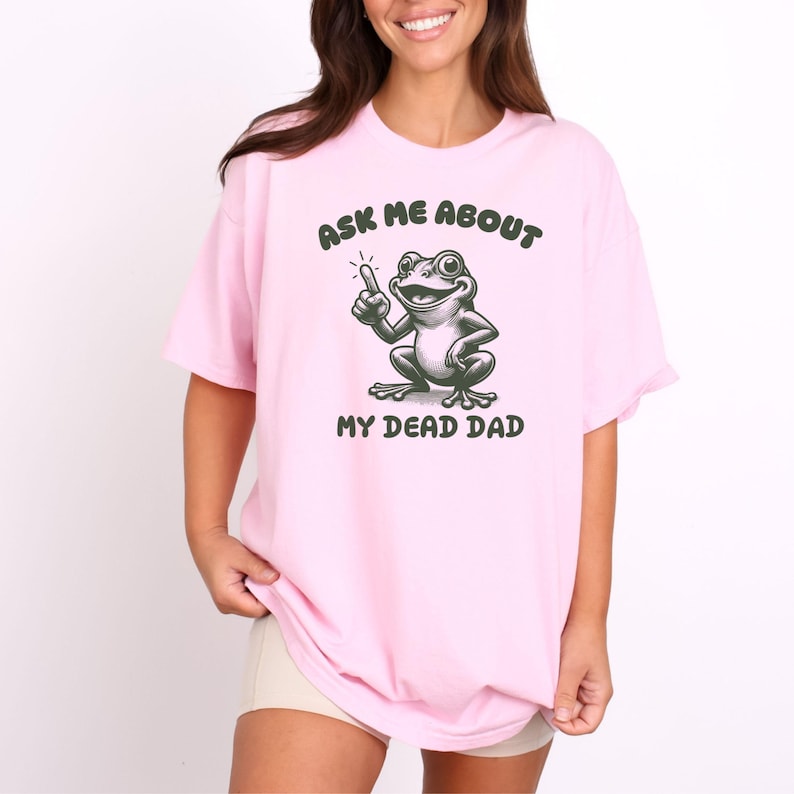 Peut inclure: T-shirt rose clair avec l'inscription "ASK ME ABOUT MY DEAD DAD" et une illustration de grenouille de dessin anim&eacute;. La grenouille est verte et blanche, pointant vers le haut avec un doigt. Le texte est vert fonc&eacute;.