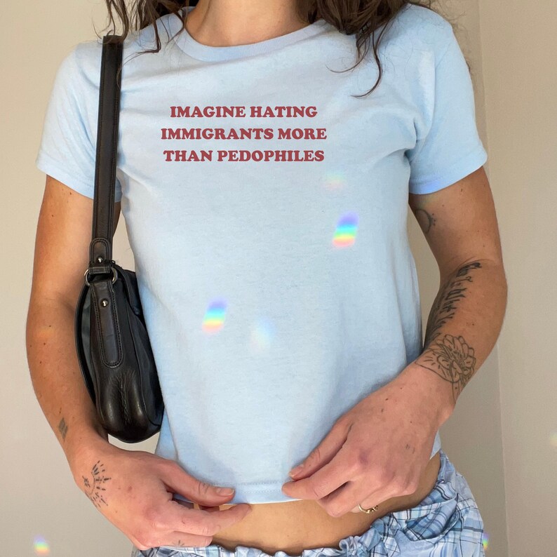 May include: Light blue t-shirt with the text "IMAGINE HATING IMMIGRANTS MORE THAN PEDOPHILES" in red. The shirt has short sleeves and a round neckline. A black purse hangs from the shoulder.