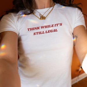 Puede incluir: Camiseta blanca con texto rojo que dice "Think while it's still legal."