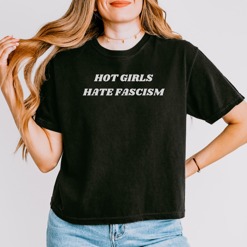 Puede incluir: Camiseta negra con el texto blanco "HOT GIRLS HATE FASCISM". La camiseta tiene cuello redondo y mangas cortas. La modelo lleva vaqueros azules.