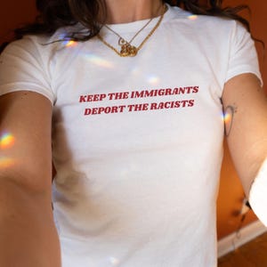 May include: White t-shirt with the red text "KEEP THE IMMIGRANTS DEPORT THE RACISTS". The shirt has short sleeves and a crew neck. A gold necklace is visible.