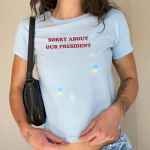 May include: A light blue t-shirt with the text "Sorry About Our President" printed in red.