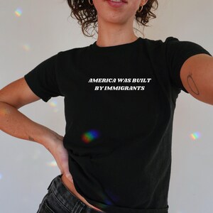 Black Baby Tee With White Text "America Was Built By Immigrants". A Political Activist And Pro Immigration Shirt For Women.