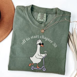 May include: An olive green t-shirt featuring a graphic of a white goose wearing a towel on its head and sunglasses, riding a purple scooter while holding a martini glass. The text above the goose reads, off to start chapter 30.