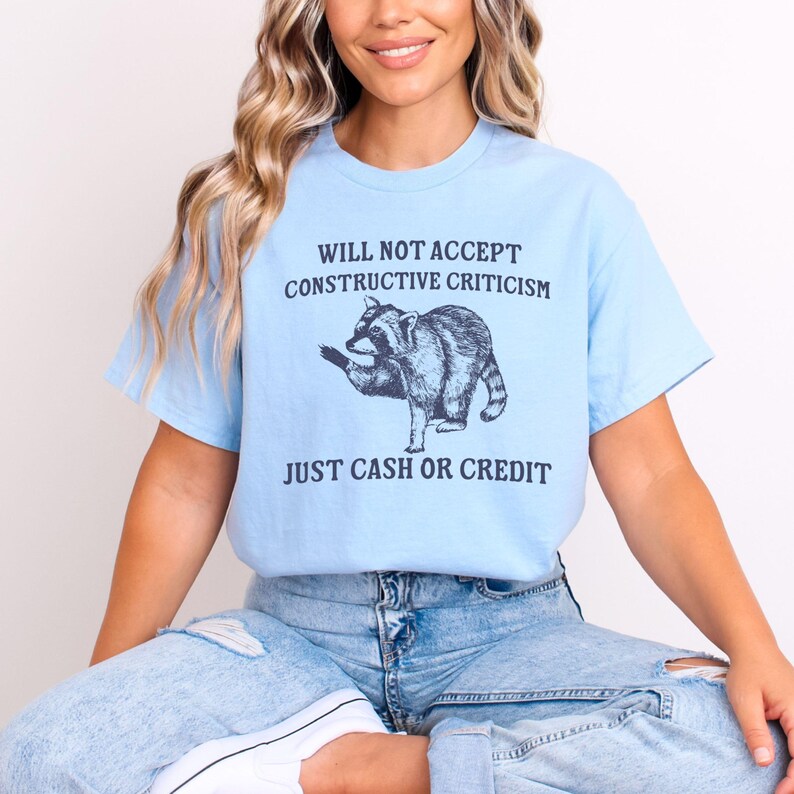 May include: A light blue t-shirt with a raccoon graphic and the text "Will not accept constructive criticism. Just cash or credit."