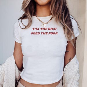 以下が含まれることがあります： 「TAX THE RICH FEED THE POOR」の赤い文字がプリントされた、クリーム色の半袖Tシャツ。長いウェーブのかかった髪と金のネックレスをつけた人が着ています。