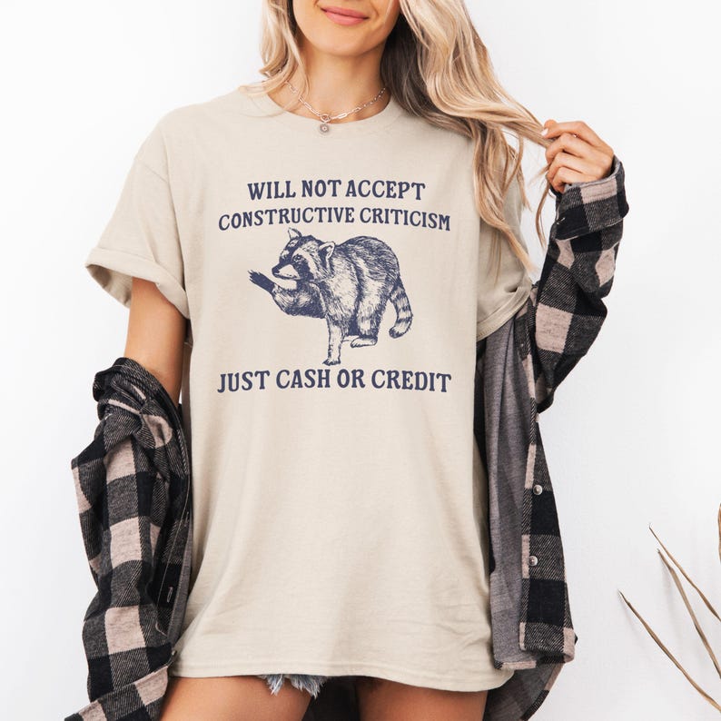 May include: A beige t-shirt with a raccoon graphic and the text "WILL NOT ACCEPT CONSTRUCTIVE CRITICISM JUST CASH OR CREDIT".
