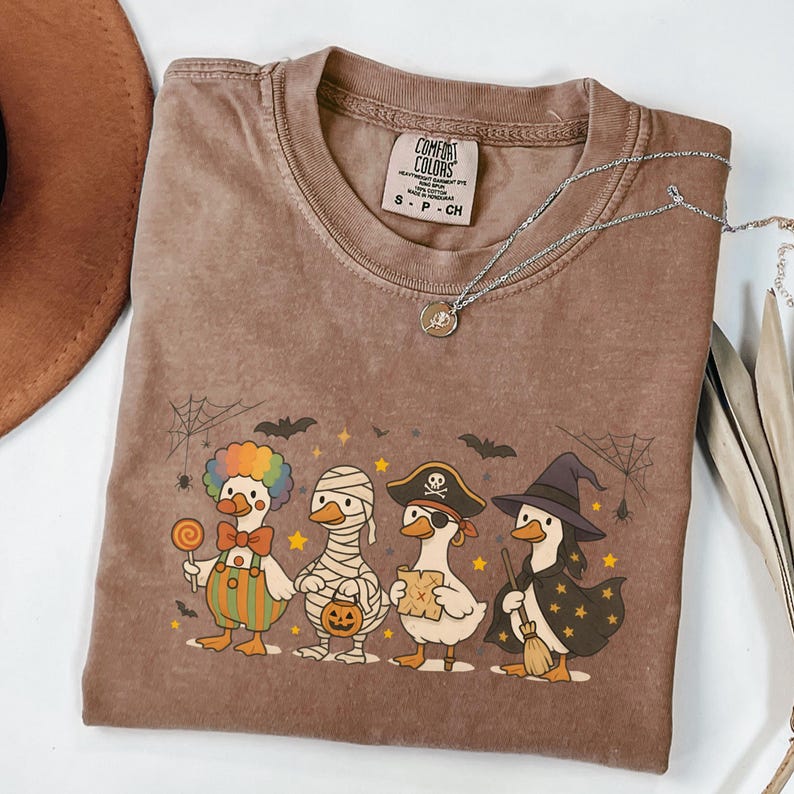 May include: Brown Comfort Colors t-shirt featuring a Halloween-themed graphic of four ducks dressed as a clown, mummy, pirate, and witch. The shirt has a crew neck and short sleeves. A silver necklace with a pendant is draped across the shirt.