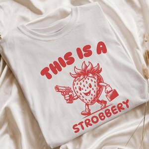 May include: White t-shirt with a red graphic of a smiling strawberry holding a gun and a briefcase. The text "This is a Strobbbery" is printed in red below the strawberry.