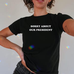 Black Y2K baby tee with white text that reads 'Sorry About Our President.' Anti-MAGA shirt, feminist baby tee, progressive liberal shirt for women.