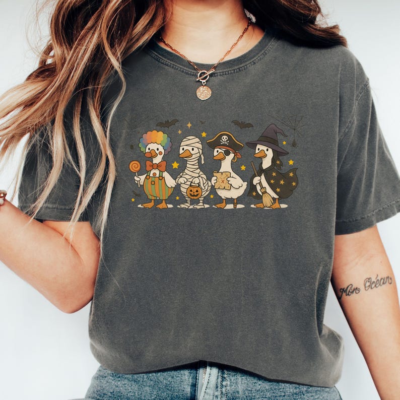 May include: Dark gray t-shirt featuring a Halloween-themed graphic. The design showcases four cartoon ducks dressed as a clown, mummy, pirate, and witch. The ducks are standing in a row with bats and stars above them. The shirt is made of a soft material.