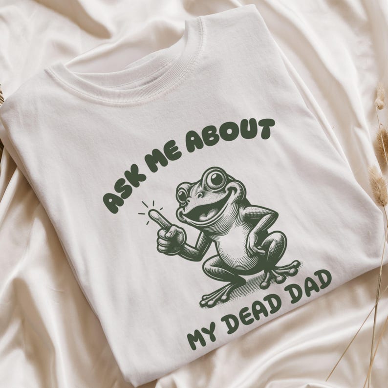 White T shirt featuring bold text that reads 'Ask Me About My Dead Dad' in green, accompanied by a cartoon drawing of a frog. This unhinged meme shirt is ideal for fans of morbid humor and the Dead Dad Club