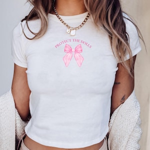 May include: White cropped t-shirt with a pink bow and the text "PROTECT THE DOLLS" printed on the front.