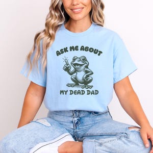 Peut inclure: T-shirt bleu clair avec le texte "ASK ME ABOUT" au-dessus d'une grenouille de dessin anim&eacute; pointant vers le haut. En dessous de la grenouille, le texte "MY DEAD DAD". La grenouille est verte et blanche.