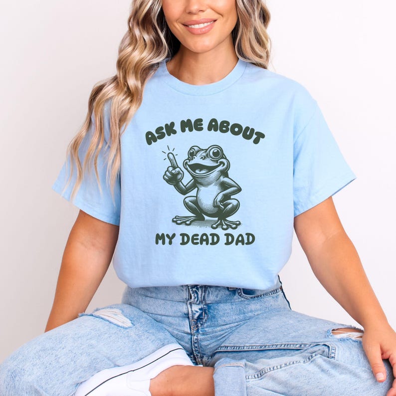 Peut inclure: T-shirt bleu clair avec le texte "ASK ME ABOUT" au-dessus d'une grenouille de dessin anim&eacute; pointant vers le haut. En dessous de la grenouille, le texte "MY DEAD DAD". La grenouille est verte et blanche.