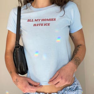May include: A light blue t-shirt with the text "ALL MY HOMIES HATE ICE" in red lettering. The shirt has short sleeves and a crew neck. A black shoulder bag is visible on the left side.