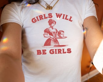 May include: White t-shirt with a red graphic print of a woman holding a platter with a man's head on it. The text "Girls Will Be Girls" is printed below the graphic.