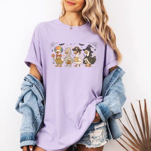May include: A light purple t-shirt featuring a Halloween-themed graphic. The graphic depicts four cartoon ducks dressed in Halloween costumes: a clown, a mummy, a pirate, and a witch. The design includes cobwebs, bats, and stars.