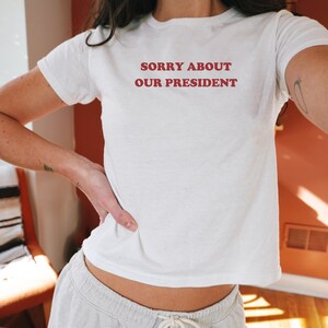 May include: White t-shirt with the text "SORRY ABOUT OUR PRESIDENT" printed in red.