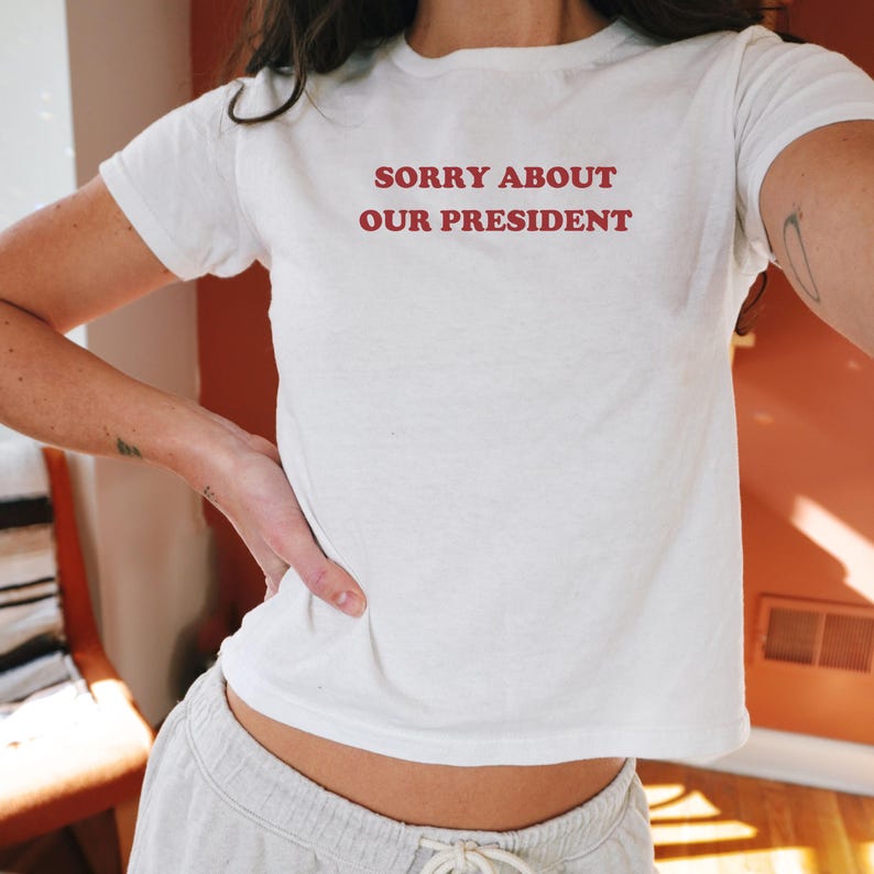 May include: White t-shirt with the text "SORRY ABOUT OUR PRESIDENT" printed in red.