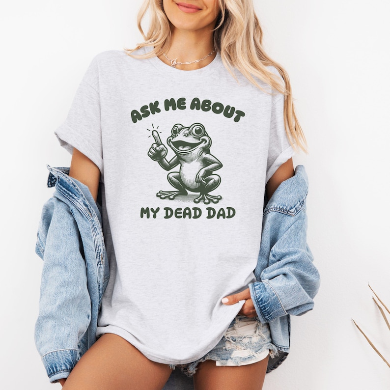 Peut inclure: T-shirt gris clair avec l'inscription "ASK ME ABOUT MY DEAD DAD" et une grenouille de dessin anim&eacute;. La grenouille verte pointe vers le haut. Le t-shirt est port&eacute; avec une veste en jean.