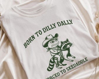 May include: White t-shirt with a green graphic of a frog in a cowboy hat riding a raccoon. The text "BORN TO DILLY DALLY" arches above the image, and "FORCED TO SKEDADDLE" is below.