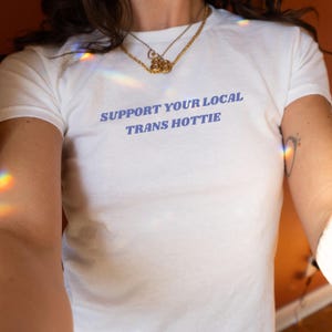 以下が含まれることがあります： 紫色の文字で「SUPPORT YOUR LOCAL TRANS HOTTIE」とプリントされた白いTシャツです。