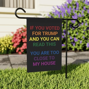 May include: A black garden flag with the text "IF YOU VOTED FOR TRUMP AND YOU CAN READ THIS YOU ARE TOO CLOSE TO MY HOUSE" in rainbow colours. The flag is displayed on a black metal stand in a garden setting with colourful flowers.