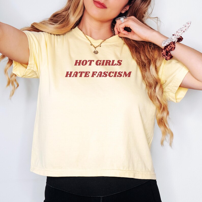 Puede incluir: Un top corto amarillo claro con el texto rojo "HOT GIRLS HATE FASCISM". La camiseta tiene mangas cortas y est&aacute; hecha de un material suave. La persona lleva un collar dorado y una goma para el pelo.