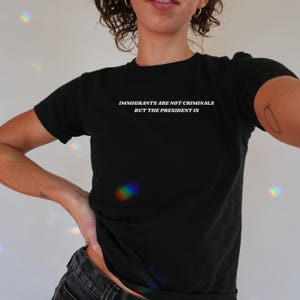 K&ouml;nnte beinhalten: Schwarzes T-Shirt mit Rundhalsausschnitt und wei&szlig;em Text: "IMMIGRANTS ARE NOT CRIMINALS BUT THE PRESIDENT IS". Das T-Shirt ist einfarbig schwarz und hat kurze &Auml;rmel.