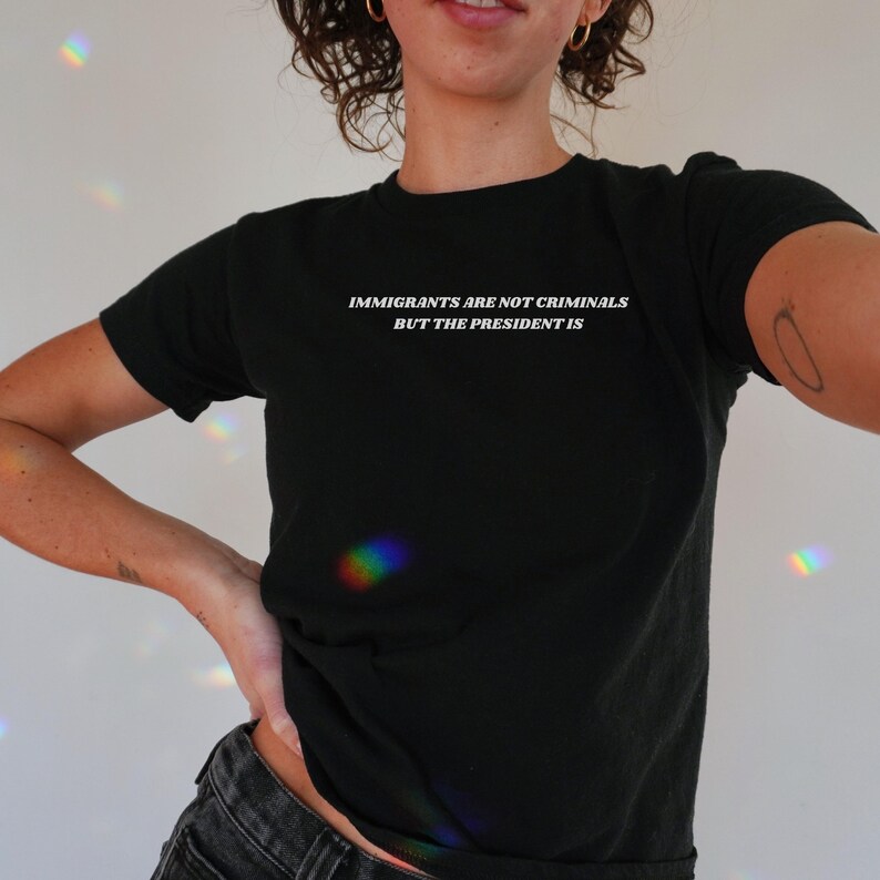 K&ouml;nnte beinhalten: Schwarzes T-Shirt mit Rundhalsausschnitt und wei&szlig;em Text: "IMMIGRANTS ARE NOT CRIMINALS BUT THE PRESIDENT IS". Das T-Shirt ist einfarbig schwarz und hat kurze &Auml;rmel.