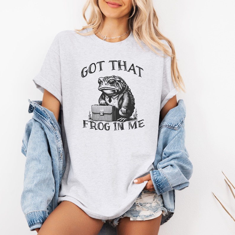Puede incluir: Una camiseta gris con un gr&aacute;fico de una rana con traje y sosteniendo un malet&iacute;n. El texto "GOT THAT FROG IN ME" est&aacute; impreso debajo de la rana.