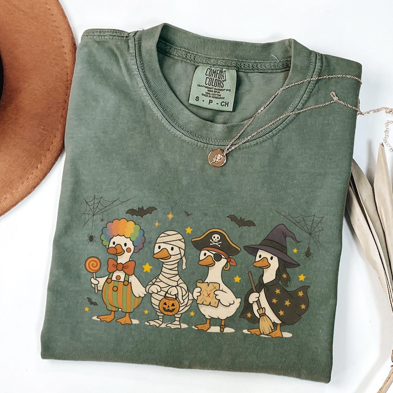 May include: Green Comfort Colors t-shirt featuring a Halloween-themed graphic of four ducks dressed as a clown, mummy, pirate, and witch. The ducks are standing in a row with bats, spiderwebs, and stars. A silver necklace is draped across the shirt.