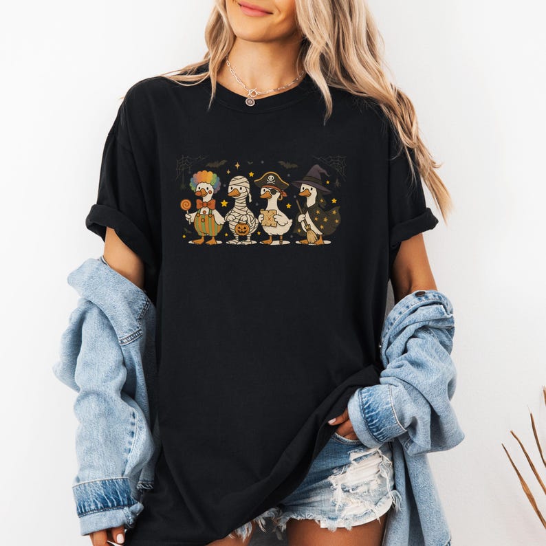 May include: Black t-shirt featuring a Halloween-themed graphic. The graphic depicts four cartoon ducks dressed in costumes: a clown, a mummy, a pirate, and a witch. The ducks are standing in a row, with stars and bats in the background.