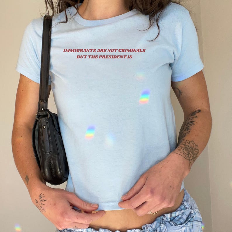 K&ouml;nnte beinhalten: Hellblaues T-Shirt mit rotem Text: "IMMIGRANTS ARE NOT CRIMINALS BUT THE PRESIDENT IS". Das Shirt hat kurze &Auml;rmel und einen Rundhalsausschnitt. Eine schwarze Umh&auml;ngetasche ist zu sehen.