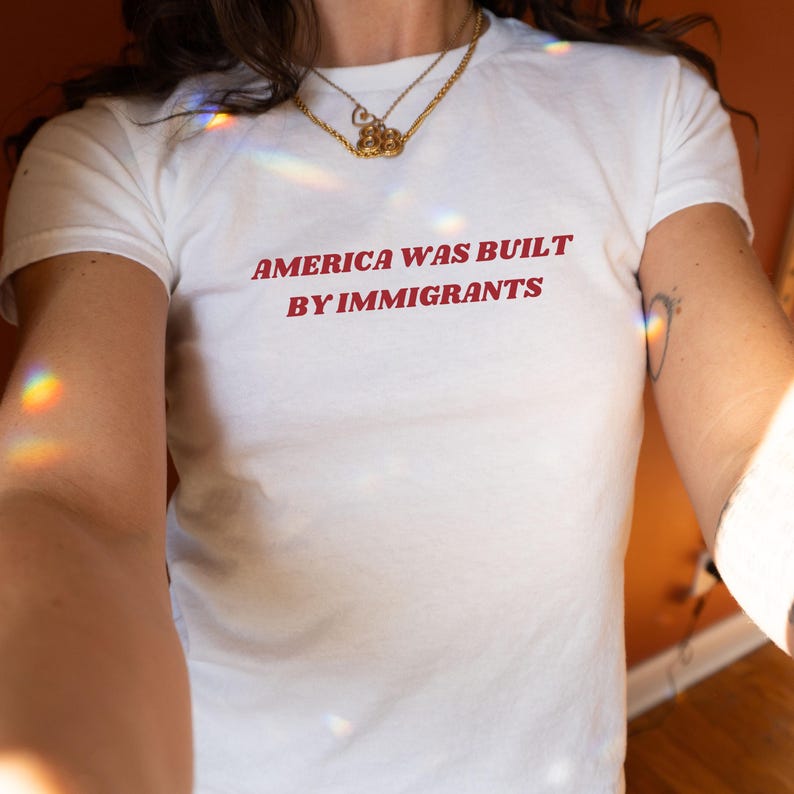 May include: White t-shirt with the red text "AMERICA WAS BUILT BY IMMIGRANTS." The shirt is short-sleeved and appears to be made of cotton. A gold necklace is visible at the neckline.
