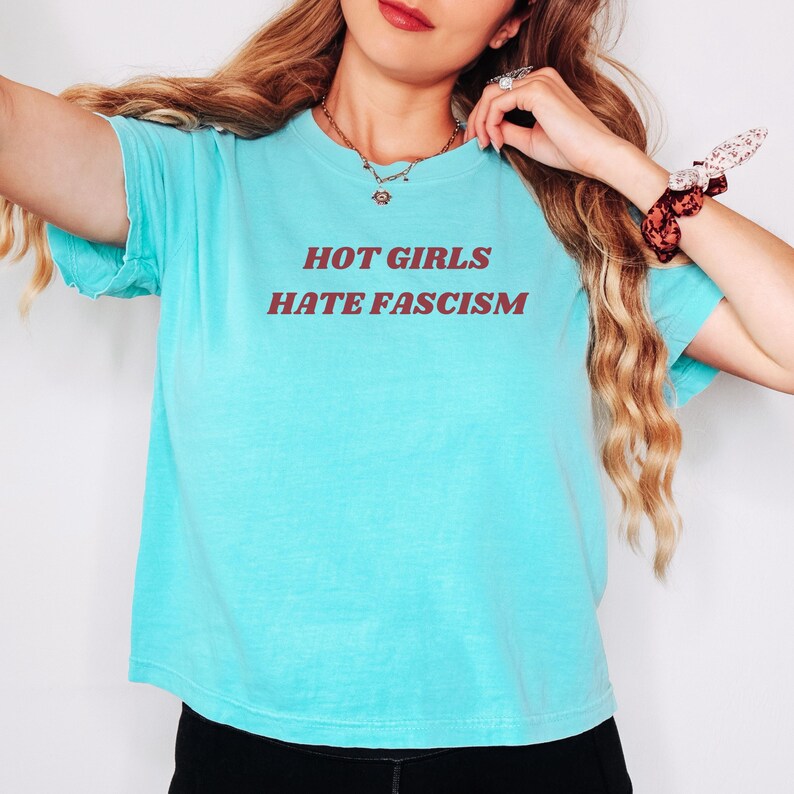 Puede incluir: Una camiseta turquesa con el texto "HOT GIRLS HATE FASCISM" en letras granates. La camiseta tiene cuello redondo y mangas cortas. La tela parece suave y ligeramente desgastada.
