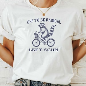 Puede incluir: Camiseta blanca con un diseño azul marino de un mapache en bicicleta y el texto "OFF TO BE RADICAL LEFT SCUM". La ilustración y el texto son azul marino.