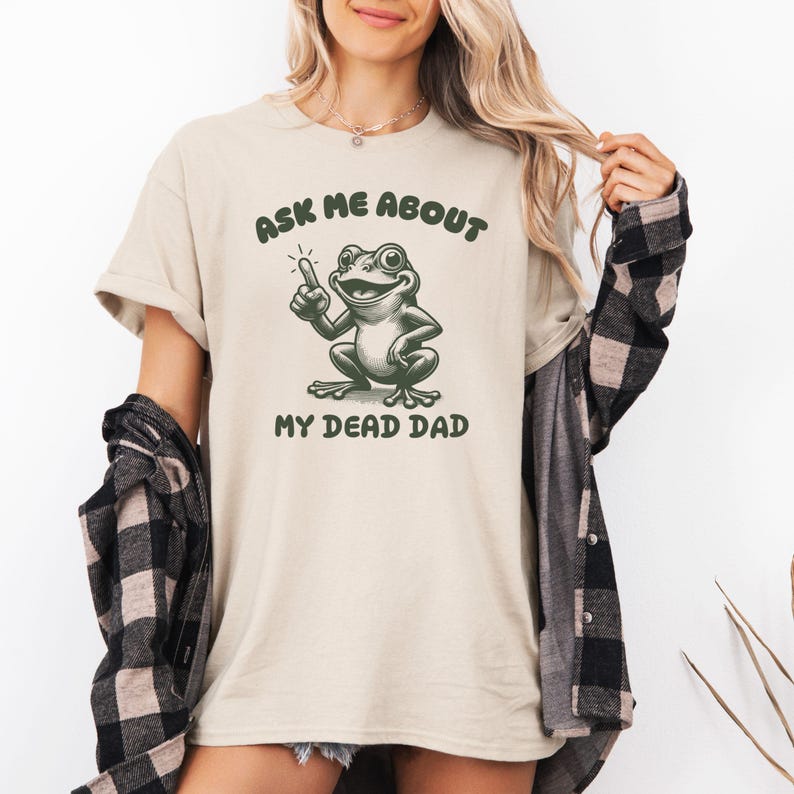 Peut inclure: T-shirt beige avec l'inscription "ASK ME ABOUT MY DEAD DAD" au-dessus et en dessous d'une illustration de grenouille verte. La grenouille pointe vers le haut. Le t-shirt est de style d&eacute;contract&eacute;.