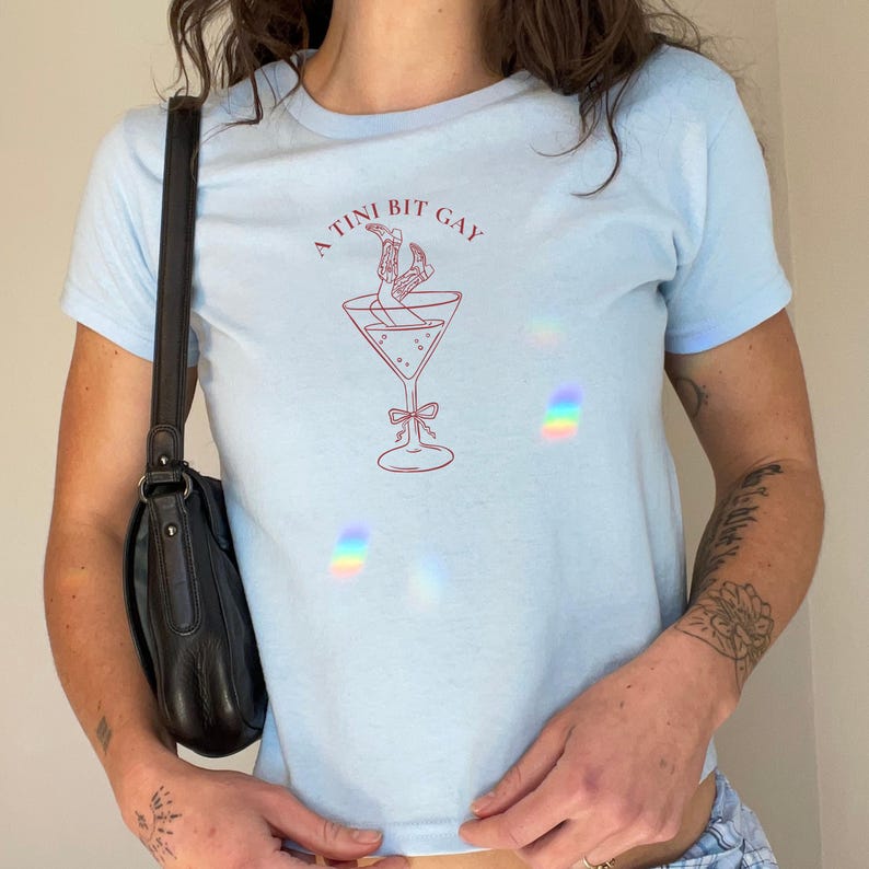 Pode incluir: Uma camiseta azul claro com um gr&aacute;fico vermelho de uma ta&ccedil;a de martini com duas pernas dentro. O texto "A TINI BIT GAY" est&aacute; acima do copo.