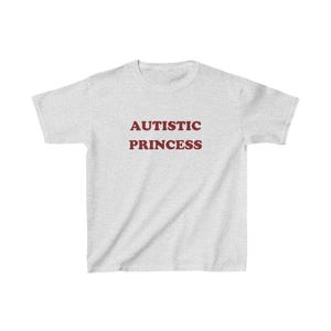 以下が含まれることがあります： 薄いグレーのTシャツに「AUTISTIC PRINCESS」という文字がマルーンでプリントされています。