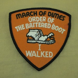Op de afbeelding: Oranje en zwarte geborduurde patch met de tekst "March of Dimes Order of the Battered Boot I Walked". Een witte cartoon-laars bevindt zich in het midden van de patch.