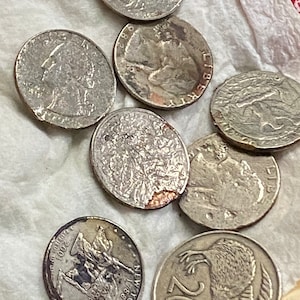 May include: A collection of eight silver coins, including US quarters and a New Zealand 20 cent coin. The coins are arranged on a white paper surface.