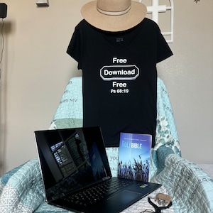 Puede incluir: Una camiseta negra con un rectángulo blanco que dice "Free Download Free Ps 68:19". La camiseta está en una cama con una computadora portátil, un libro titulado "Holy Bible" y un collar con una cruz.