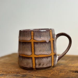 May include: A handmade ceramic mug with a brown handle and a grid pattern. The mug features a brown and tan color scheme with orange grid lines. The mug is sitting on a wooden surface.