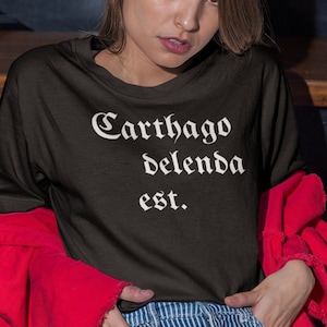 May include: A black t-shirt with the text "Carthago delenda est." in white lettering. The shirt is being worn by a person sitting on wooden stairs.