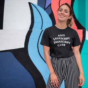 May include: A woman wearing a black t-shirt with the text "ANTI JAVASCRIPT JAVASCRIPT CLUB" in white. She is standing in front of a colorful wall with abstract shapes.