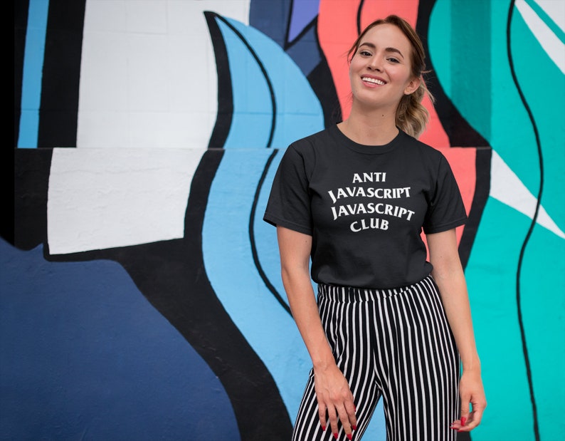 May include: A woman wearing a black t-shirt with the text "ANTI JAVASCRIPT JAVASCRIPT CLUB" in white. She is standing in front of a colorful wall with abstract shapes.